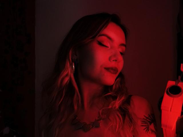 AlanisBlanc mit rot beleuchtetem Gesicht, welligem blondem Haar, dunklem Lippenstift, geschlossenen Augen, einer Halskette, einem Tattoo auf der Schulter, die ein rotes Licht hält, mit Ohrringen, mit einem sanften Lächeln. Das rote Licht wirft einen warmen Schein..