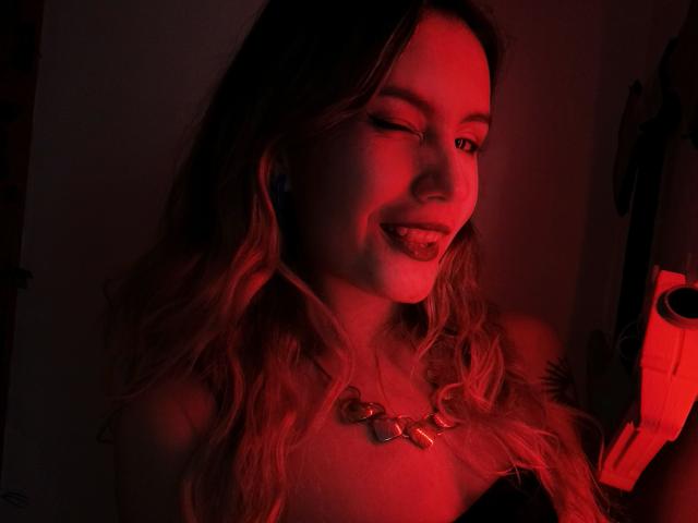 AlanisBlanc, avec ses cheveux blonds ondulés, un visage illuminé de rouge, la langue tirée, portant un collier, tenant un objet rouge, souriant légèrement, ses seins à peine visibles dans la faible lumière..