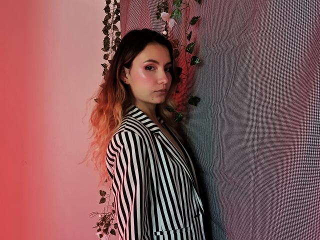 Alanis Blanc, com cabelos castanhos longos e ondulados, com pontas laranja, vestindo um blazer listrado preto e branco, olhando por cima do ombro, em pé em frente a uma parede cinza com textura, com trepadeiras verdes penduradas..
