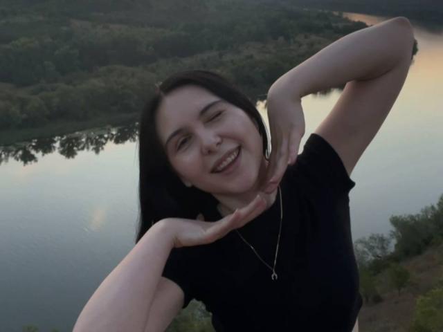 MapleUrby with black hair, smiling, wearing a black shirt, hands near neck, small necklace, pale skin, standing outdoors at dusk, reflection in water, slightly blurry.