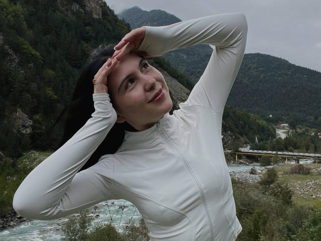 MapleUrby with hands on head, wearing a white long-sleeve top, smiling, standing by a river, forested mountains in background, overcast sky, her tits slightly visible under the top.