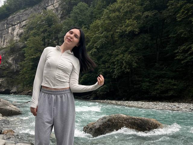 MapleUrby stands by a river, wearing a white long-sleeve top and gray sweatpants, smiling with her dark hair blowing, showing her small tits and slim, cute ass.