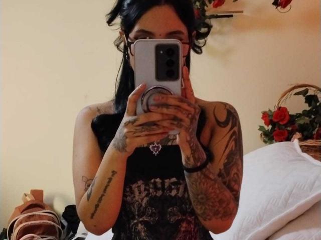 GabyMathew with tattoos, holding phone in front of face, black sleeveless top, dark hair in pigtails, taking mirror selfie, visible arm tattoos, white pillow in background.