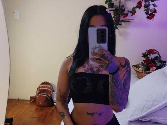 Gaby Mathew mit langen, schwarzen Haaren, trägt ein schwarzes, netzförmiges BH-Top und zeigt Tattoos an Armen und Brust. Sie macht ein Selfie vor einem Spiegel mit ihrem Handy und zeigt dabei ihre Brüste und ihren tätowierten Bauch..
