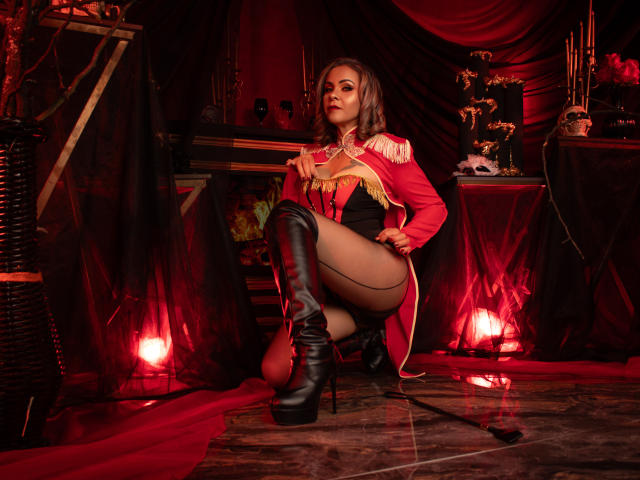 VictoriiaGreys in a red military-style jacket with gold epaulettes, black stockings, and thigh-high black boots, squatting, holding a whip, with red lighting and dark drapes.
