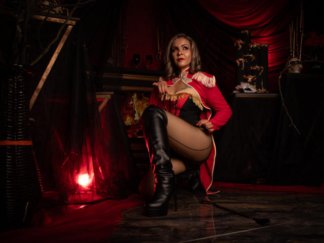 VictoriiaGreys squats in a dark, red-lit room wearing a red cape, black corset, and thigh-high boots, with her ass and tits prominently displayed, holding a whip.