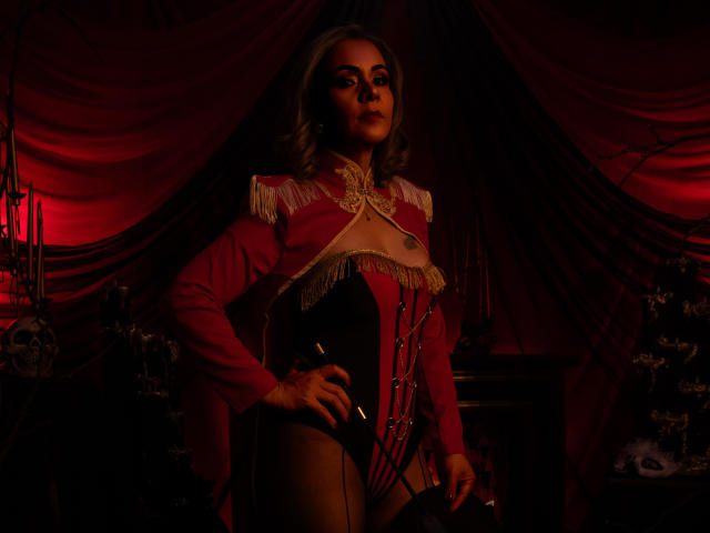 VictoriiaGreys draagt een donkerrode top met franjes en lange mouwen, met kantdetails, een zwarte corset en kniekousen. Ze staat met één hand op haar heup, met rood licht..
