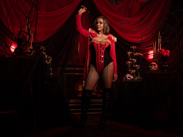 VictoriiaGreys in un corsetto rosso con pannelli neri trasparenti, stivali neri alti fino alla coscia, che tiene un anello, in piedi in una stanza buia, drappeggiata di rosso, con teschi e candele..