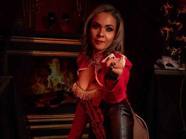 VictoriiaGreys with grey hair, red jacket, gold fringes, and black leather pants, pointing, showing off her tits, in a dimly lit, sexy, Victorian-style room.