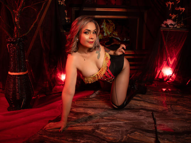 VictoriiaGreys on all fours, wearing a red and black fringed corset, black panties, and boots, with visible tits, tattoo on left shoulder, red lights, and a sexy expression.