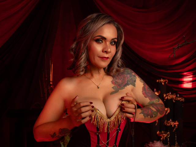 VictoriiaGreys, met grijs krullend haar, grote borsten, tatoeages op de armen, draagt een rode corset met gouden franjes, haar handen bedekken haar tepels, rode ogen, donkerrode lipstick, in een donkere kamer..
