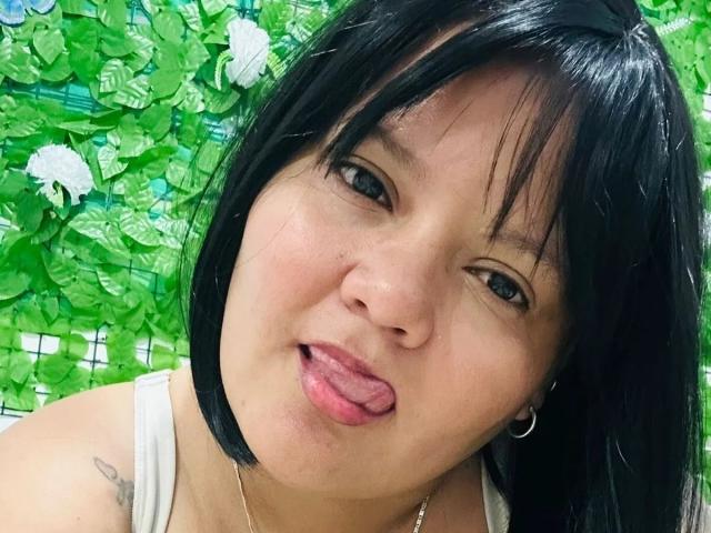 CamilaBacyc with black hair, sticking out her tongue, green eyes, small hoop earring, tattoo on left shoulder, white tank top, and a cute, playful expression.
