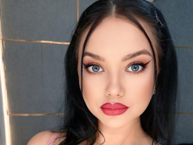 JessyQuinn has bright blue eyes, dark straight hair, and red lipstick. Her makeup includes thick eyeliner and she's wearing a pink top. Her face is very pretty.