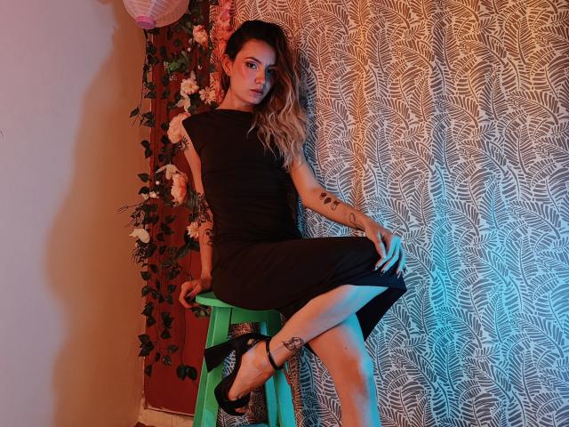 AlanisBlanc, avec de longs cheveux blonds, vêtue d'une robe noire, est assise sur un tabouret vert, montrant sa jambe tatouée et ses talons noirs, devant un rideau à motifs, dans une lumière bleu clair et rose..