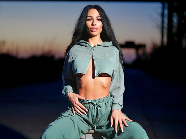 CharlotteAria with long black hair, green crop hoodie, and green pants, sitting, showing her toned abs, hands on her pants, sunset in the background, soft light on her face.