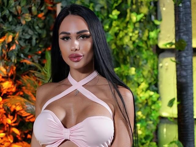CharlotteAria with long black hair, wearing a pink, criss-cross, strapless top that shows her big, shiny, round tits. She's looking at the camera with pouty, pink lips.