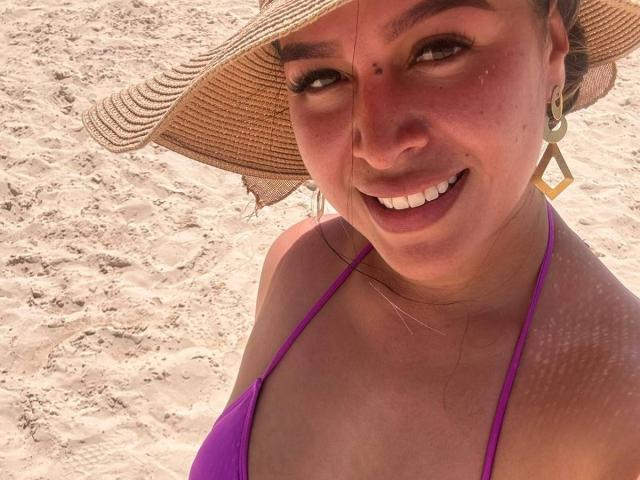 LiaDiane, com a pele bronzeada, sorrindo, vestindo um top de biquíni roxo, um chapéu de palha e brincos de ouro, tomando sol em uma praia de areia, mostrando seus seios e uma pequena marca de beleza..