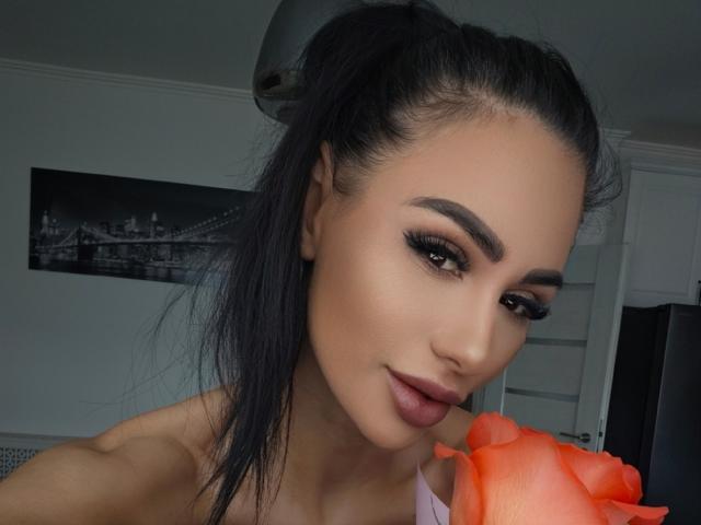 CharlotteAria with dark hair in a ponytail, thick eyebrows, long eyelashes, full lips, holding an orange rose, wearing a pink top, smiling slightly, in a selfie.