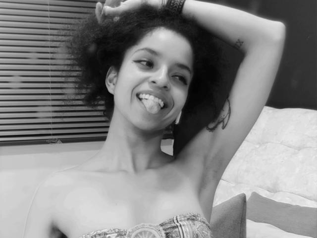 ErikaLara with dark curly hair, sticking out her tongue, showing off small tits with one arm raised, a tattoo on her arm, and a strapless patterned top. Black-and-white photo.