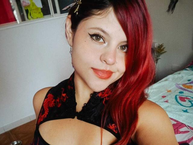 Sophie Hill, avec ses cheveux roux, son haut noir à lacets, son anneau de nez, son eye-liner et son rouge à lèvres orange, sourit à l'appareil photo. Ses seins sont partiellement visibles à travers le haut..