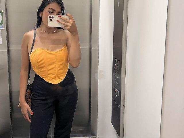 AmeliRoux, avec ses cheveux noirs mouillés, portant un haut jaune sans bretelles et un pantalon noir, tenant un téléphone contre son visage, montrant ses petites seins et ses fesses fines dans un miroir..