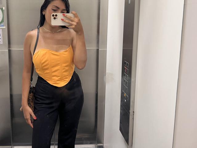 AmeliRoux with tan lines, wearing a yellow strapless top, black high-waisted pants, holding a phone covering her face, black hair, necklace, and a leopard print bag.
