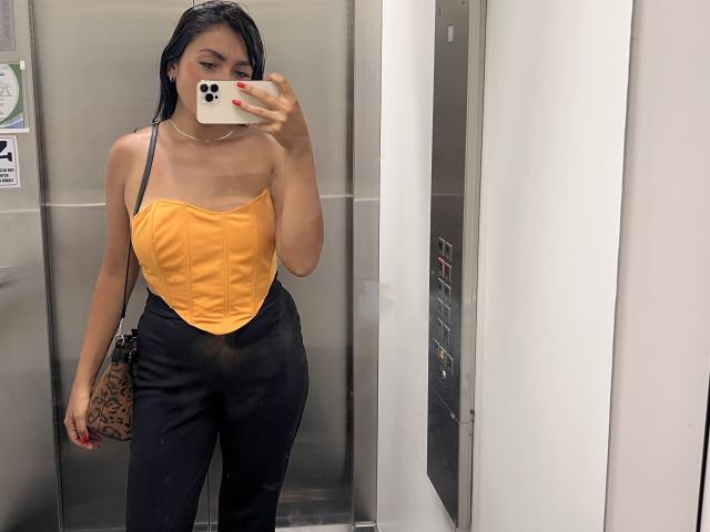 AmeliRoux, avec ses cheveux noirs mouillés, un haut jaune sans bretelles, un pantalon noir taille haute, un sac à motifs léopard, des ongles rouges, tenant un iPhone, un selfie devant un miroir, debout dans un ascenseur. Seins légèrement visibles..
