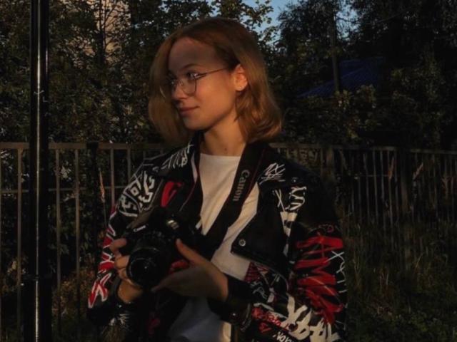 LoniTockey with glasses, holding a camera, wearing a black jacket with red and white patterns, standing outside, backlit by evening sunlight, looking to the side, arms slightly forward.