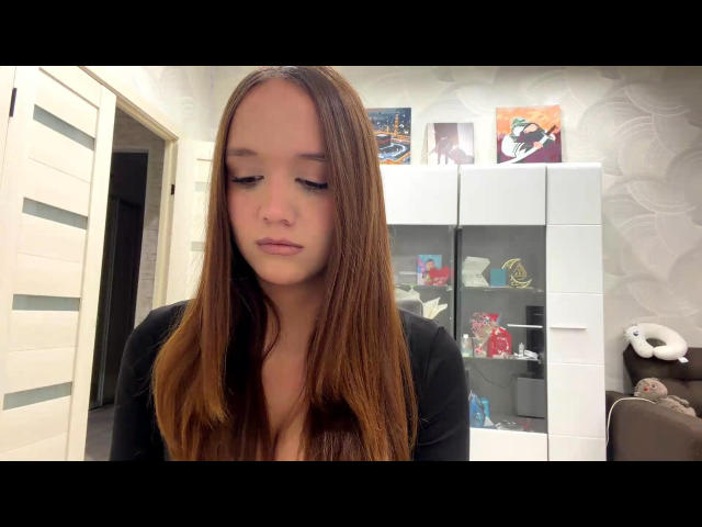 NuPastiva with long brown hair, small tits, and a serious face, wearing a black top, in a room with a white cabinet and stuffed animal.
