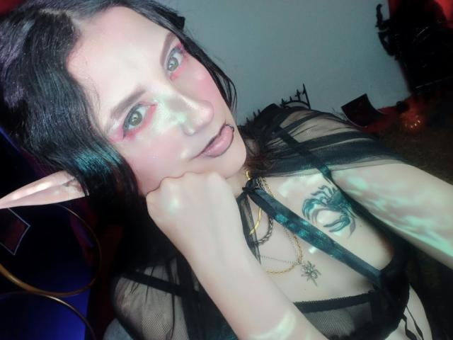 MaryCruzz with pale skin, black hair, elf ears, pink eye makeup, black sheer top, tongue out, hand on cheek, tattoo on right chest, gold necklace, in a dimly lit room.