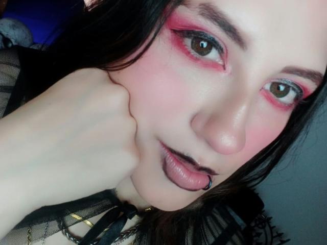 MaryCruzz's face with pink eye makeup, black lipstick, nose ring, and black mesh top. She rests her chin on her hand, showing pale skin and dark brown eyes.