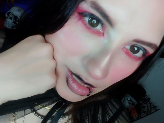 MaryCruzz with pink eyeshadow, black lipstick, and a pierced tongue, resting her cheek on her hand, showing off her pale skin and brown eyes in a selfie.