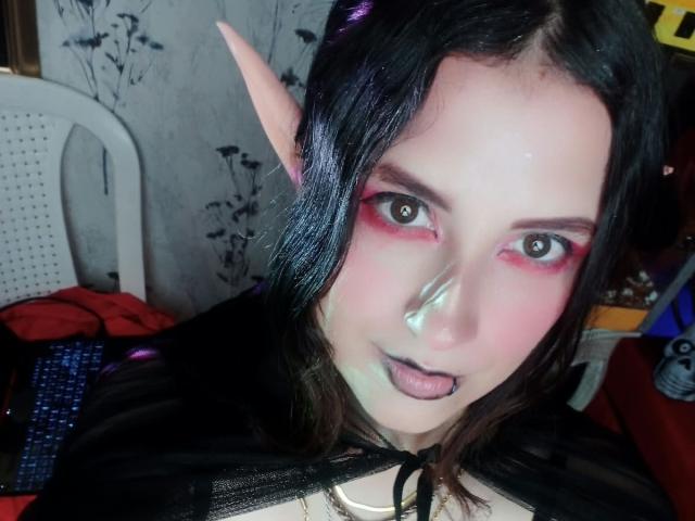 MaryCruzz has pale skin, black lipstick, pink eye makeup, elf ears, black hair, and a black cape. She's sticking out her tongue with a cute, playful expression.