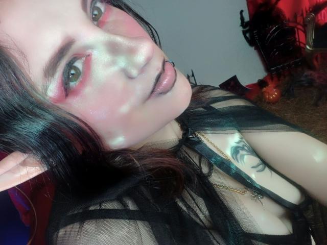 MaryCruzz with pale skin, pink eye makeup, black mesh top, pouty lips, pointed elf ears, black hair, tattoo on chest, and white makeup on face. Boobs visible under mesh.