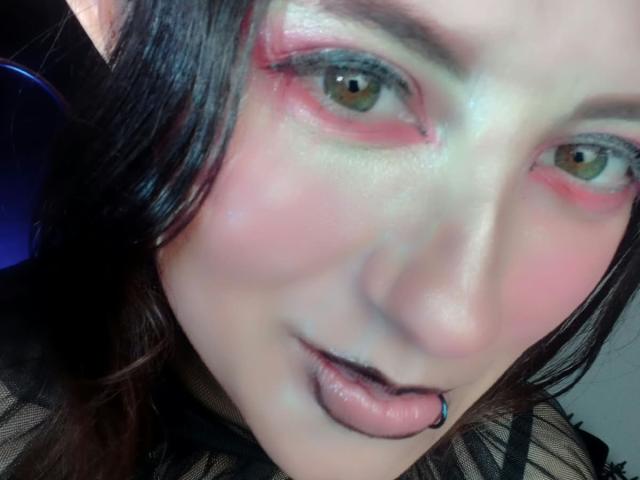 MaryCruzz's face, pale skin, pink eyeshadow, brown eyes, black lipstick, tongue piercing, dark hair, slight blush, wearing black mesh top, looking at camera with slight smile.