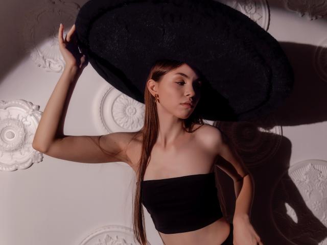 AgataRyili with long brown hair, wearing a black tube top and a huge black floppy hat, holding it above her head, showing her slim, pale, and pretty face.