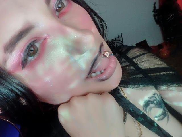 MaryCruzz with pink eye makeup, black lipstick, septum piercing, and black sheer top, holding her chin, showing a tattoo on her shoulder, in a low-light, shiny-skinned selfie.