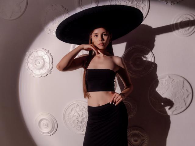 AgataRyili stands with a hand on her chin, wearing a black strapless top and wide hat, black skirt, long brown hair, light skin, and a belly button piercing.