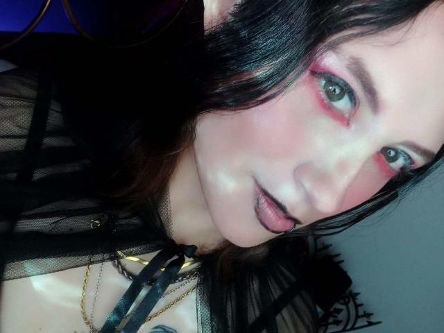 MaryCruzz with black hair, pink eyeshadow, black lipstick, and a sheer black top, showing a slight tongue-out expression, looking directly at the camera.