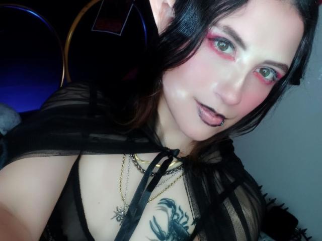 MaryCruzz has pink eye makeup, black lipstick, and a black sheer top, showing her tits with a tattoo above. A man's head is partially behind her.