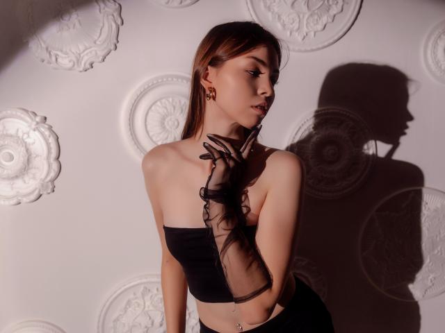 AgataRyili with long brown hair, wearing black strapless top and sheer glove, touching her chin, shadow on wall, showing her small tits and belly button piercing.