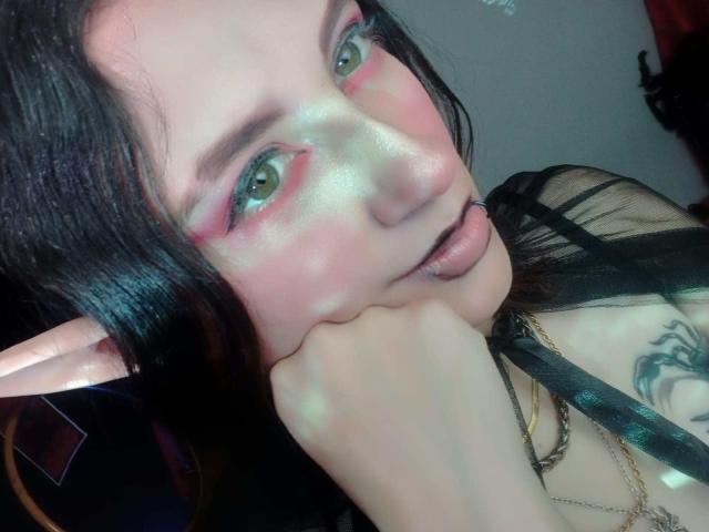 MaryCruzz has pale skin, green eyes, black lipstick, pink eyeshadow, and black hair. She's biting her finger with elf ears, a tattoo on her shoulder, and a black strap.