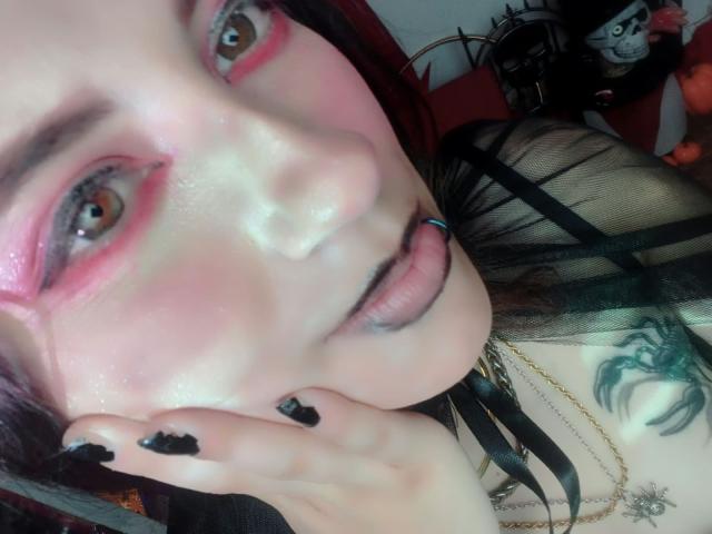 MaryCruzz's face with pink eye makeup, black lipstick, black nail polish, blue tongue piercing, and green spider tattoo on chest, wearing black mesh top and gold necklace.