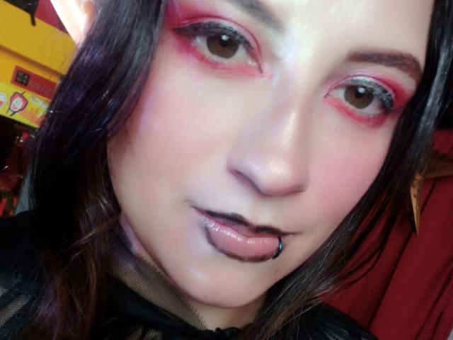 MaryCruzz has pink eye makeup, black lipstick with a blue piercing, and a pale complexion. Her dark hair is partially visible, and she's wearing a black mesh top.