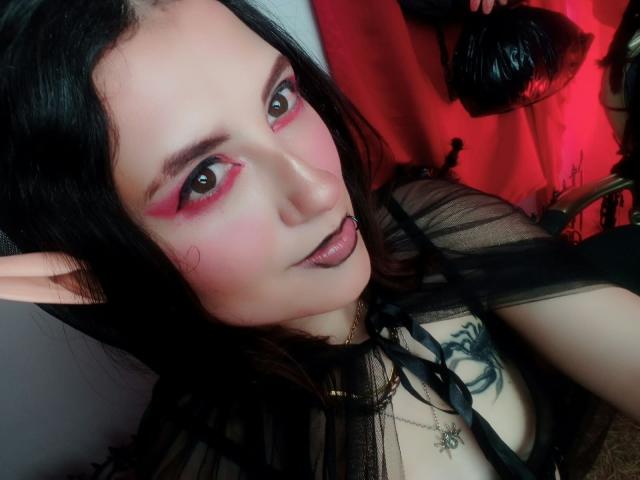 MaryCruzz with pink eye makeup, black lipstick, and elf ears, wearing a sheer black top, showing a tattoo on her chest, holding a black flogger.