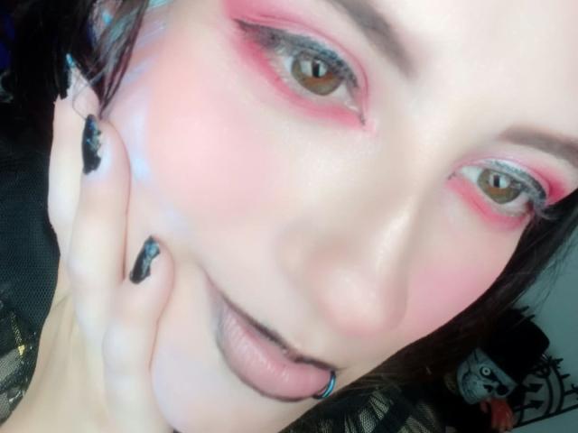 MaryCruzz has pink eyeshadow, black nail polish, and a pierced bottom lip. She's holding her cheek with her hand, showing off her glossy skin and brown eyes.
