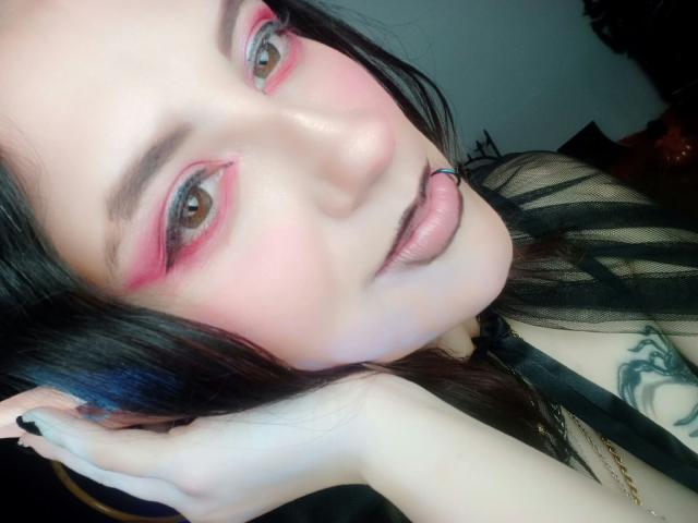 MaryCruzz has pink eyeshadow, black lipstick, a blue lip ring, and black striped shirt. Her hand touches her head, showing a tattoo. She has fair skin and dark hair.