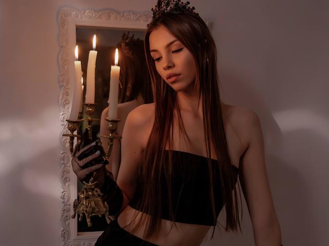 AgataRyili with long brown hair, wearing a black strapless top, black gloves, and a black tiara, standing in front of a mirror with lit candles, looking down.