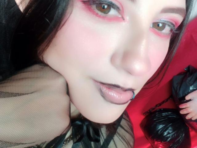 MaryCruzz's face, pink eyeshadow, black lipstick, septum piercing, pale skin, dark hair, sheer black top, red background, black bag on right.