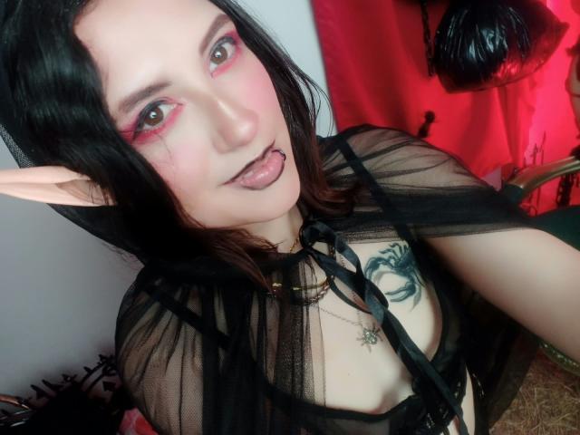 MaryCruzz with pale skin, black hair, pink eyeshadow, pointed elf ears, black sheer top, tongue out, black lip ring, tattoo on chest, red background, and black leather hanging.